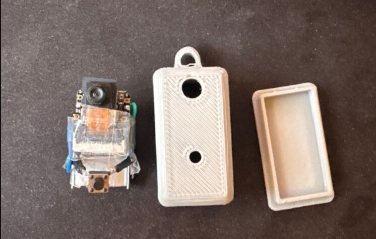 ESP32-S3 keychain camera exploded view — camera module with tactile button, 3D-printed body, translucent lid